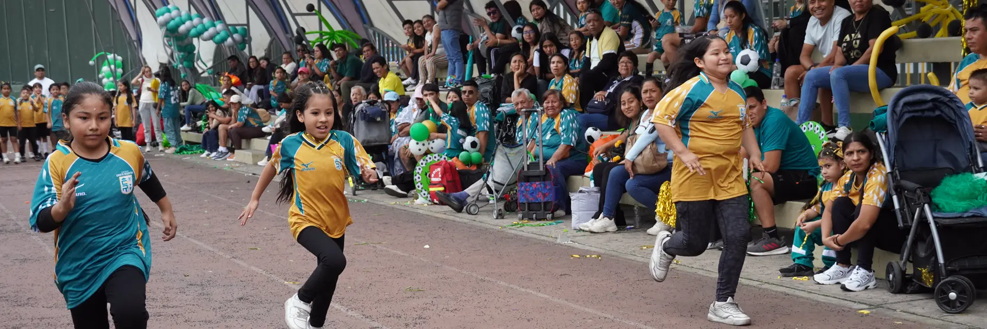 Tres estudiantes corren en la pista atlética durante las Olimpiadas Redentorinas, con las familias en la tribuna.